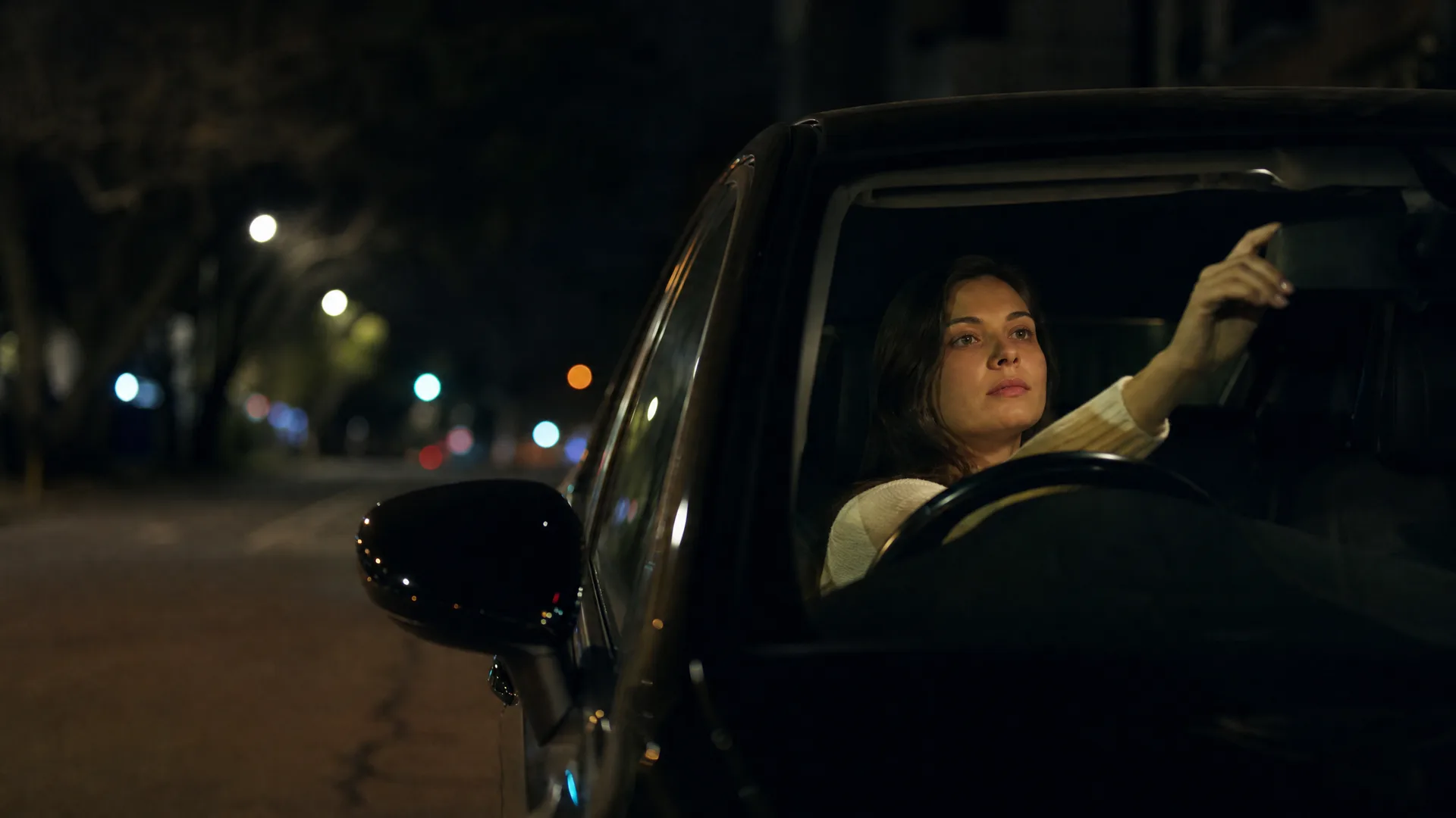 A woman sits in the driver's seat of a car at night with one hand raised toward the rear-view mirror, with blurred street lights and trees along the road behind her.